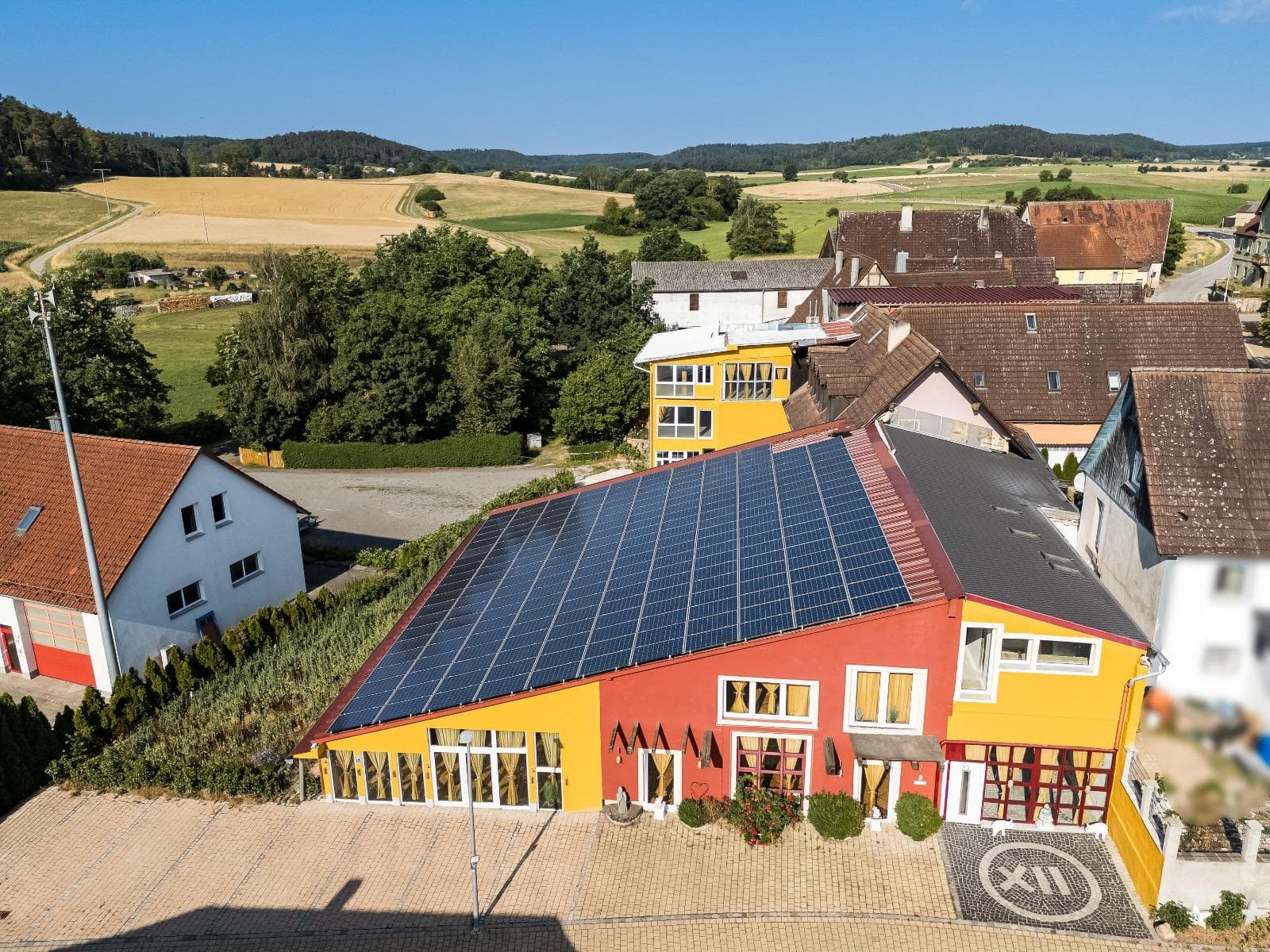 Luftaufnahme von Punkt XII – das rot-gelbe Haus mit großer Solaranlage vor Feldern und Wald